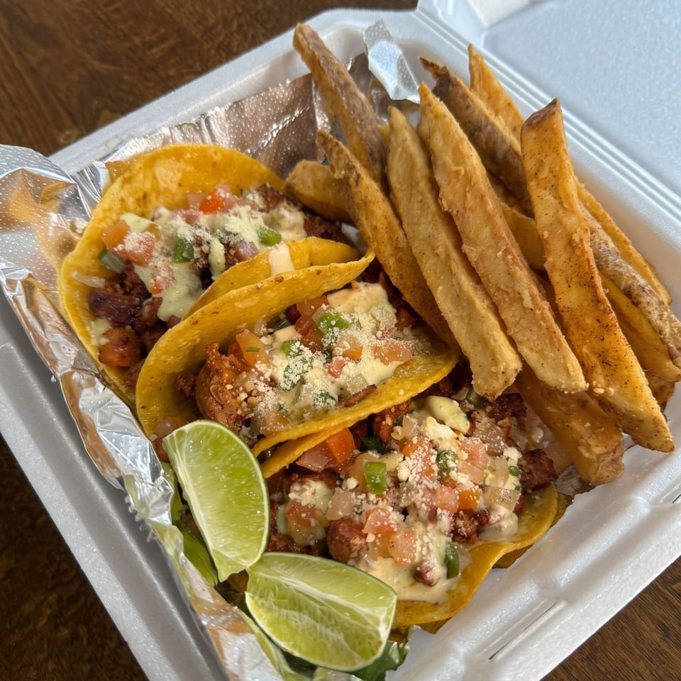 Three street tacos with lime wedges and a side of fries in a white takeout container.