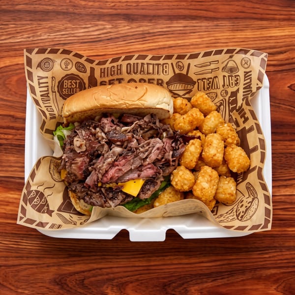 Steak sandwich with melted cheese and vegetables served alongside golden tater tots in a paper-lined takeout container.