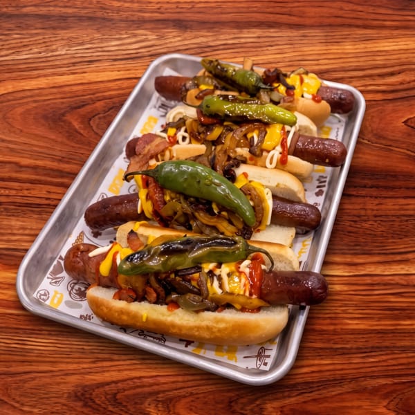 Tray of loaded hot dogs topped with grilled peppers, onions, cheese, mustard, ketchup, and mayonnaise on a wooden table.