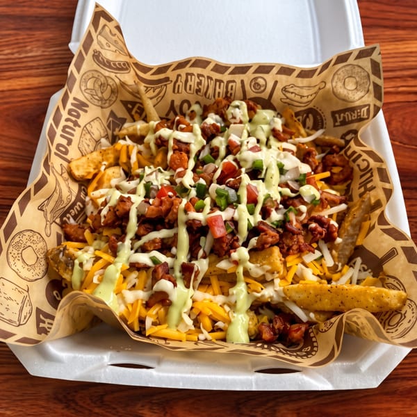 Loaded fries topped with cheese, bacon, diced vegetables, and creamy green sauce in a branded paper-lined tray.