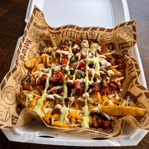 Loaded fries topped with seasoned meat, shredded cheese, diced vegetables, and creamy green and white sauces in a takeout tray.