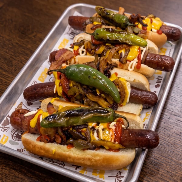 Tray of loaded hot dogs topped with grilled peppers, onions, bacon, cheese, and drizzled sauces on a wooden table.
